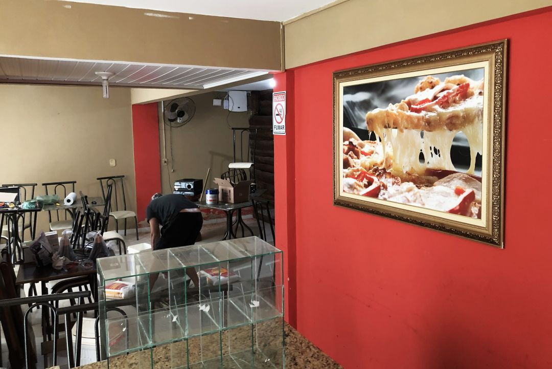Interior de estabelecimento comercial com parede vermelha ao lado, destacando um quadro grande com foto de pizza com queijo derretido. Ao fundo, mesas e um colaborador trabalhando.